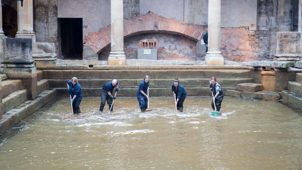 Five people in blue overalls, sweeping water in the Roman Baths