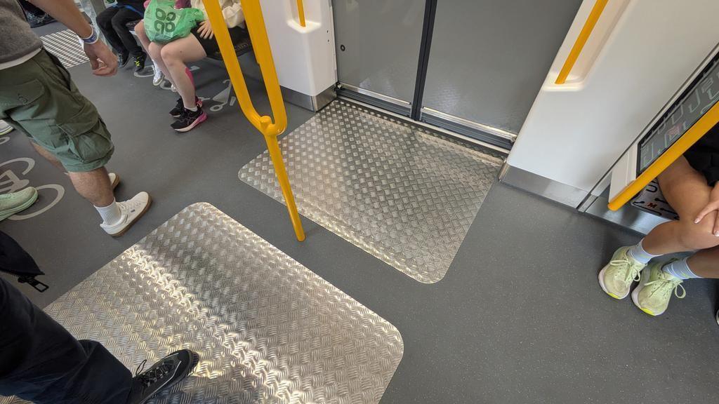 Two metal zig zag plates on each side of a train carriage door on the floor. A yellow bar rail is in the middle of them. The legs and feet of passengers standing and sitting down can be seen.