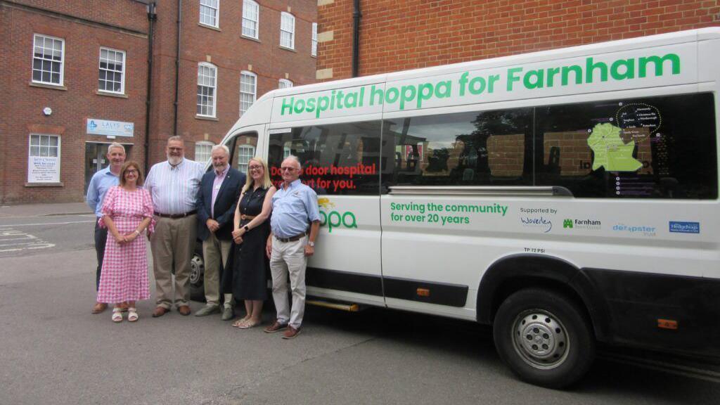 A community transport van labeled "Hospital hoppa for Farnham" is parked in front of a group of people. The van is white with green branding and text. Behind them, red-brick buildings with large windows line the street.