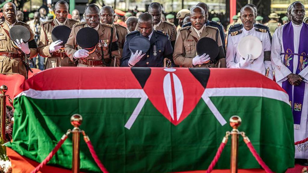 Police officers lowering their ceremonial hats in solemn tribute before a coffin draped in the Kenyan flag