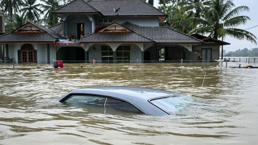 A car submerged almost all the way to the roof in murky floodwater. In the background is a house, also flooded with water at a very high level. 