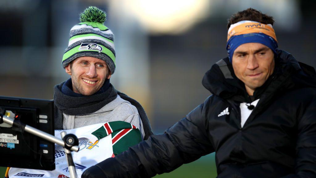 Sinfield embraces his friend Burrow at the end of a fundraising challenge. Sinfield wears his fundraising head band and a black thermal jacket, Burrow is wrapped in a woolly hat, scarf and blanket and smiles. Burrow's computer, which aided his speech, can be seen in the shot.