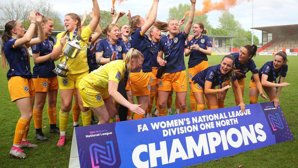 A group of about 15 players from Hull City Ladies. Most are wearing orange shirts and socks with blue tops. Two are wearing yellow shorts and tops. They are learning over a banner that reads: "FA Women's National League Division One North Champions. One of the players is holding a large silver trophy and some of the girls have their arms in the air.