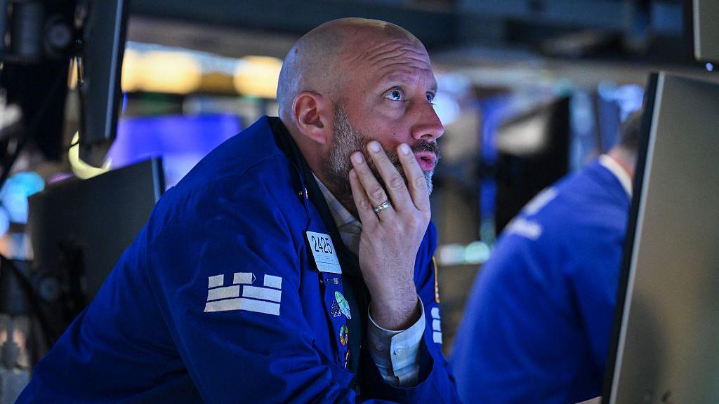A trader in a blue coat holds his chin in his hand while looking with furrowed brow at a computer on the floor of the New York Stock Exchange (NYSE) in New York City.
