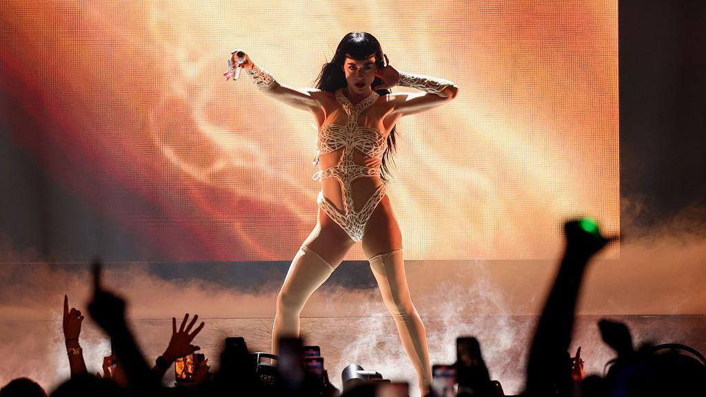 Katy Perry is on stage and is wearing a white and nude bodysuit with a lace cross over her stomach. She is holding out a microphone to a crown who have their arms and phones in the air.