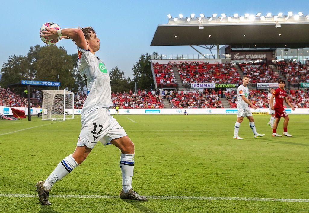 Callan Elliot in a white football strip takes a long throw-in as a couple of other footballers look on with a stadium and floodlights in the background