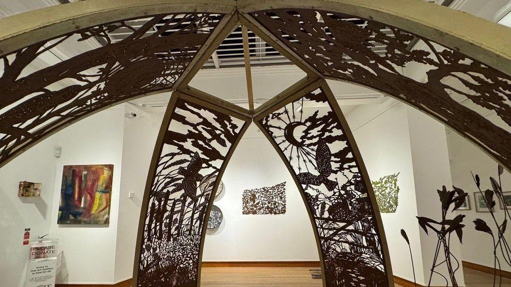 A trellis-type metal artwork that has birds, grasses and a moon sculpted into it. It is positioned in the centre of the art gallery, with other exhibits on the white walls.