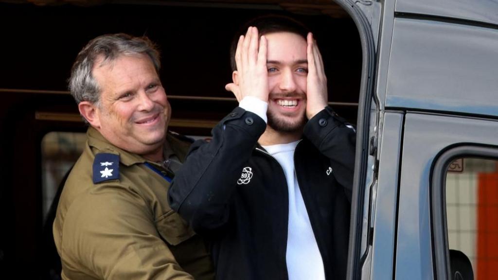 Omer Shem Tov holding his face with both hands and smiling, stood in a van with a man in uniform next to him.