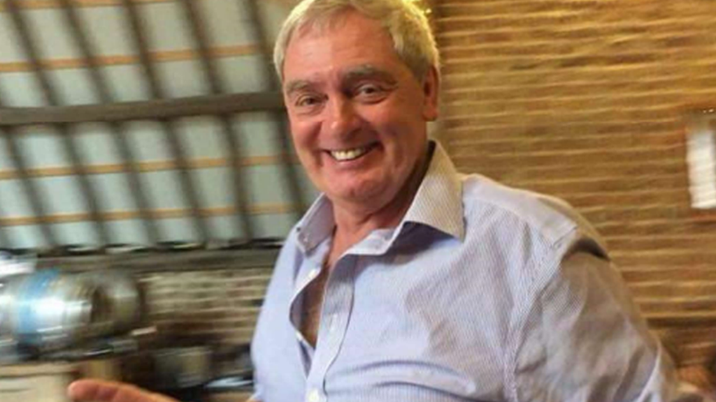 A 65 year old man smiling at a camera with a pint in his hand inside a pub