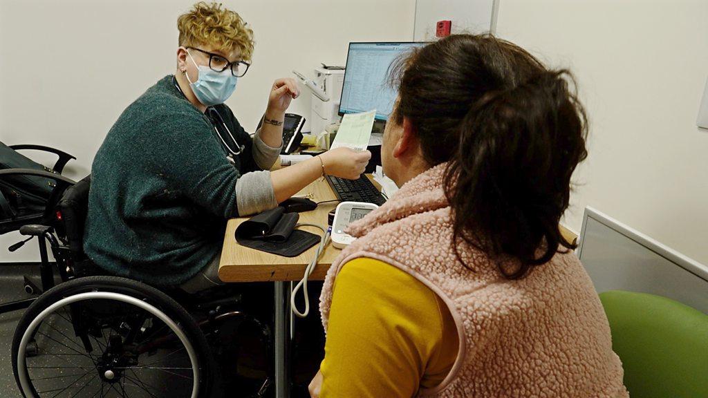 Georgie in her wheel chair in GP treatment room talking to a patient