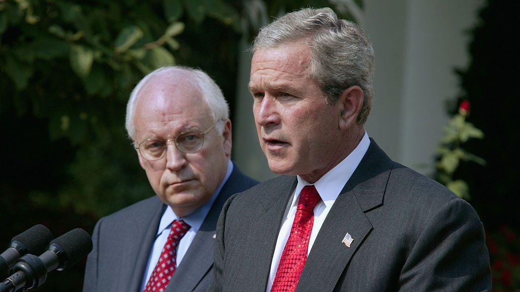 George W Bush speaks at a lectern at the White House in 2004, and Cheney, standing next to him, looks at him.