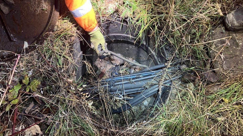 A hole in the ground containing wires. A person wearing a yellow glove is holding one of the wires as teams try to carry out repairs