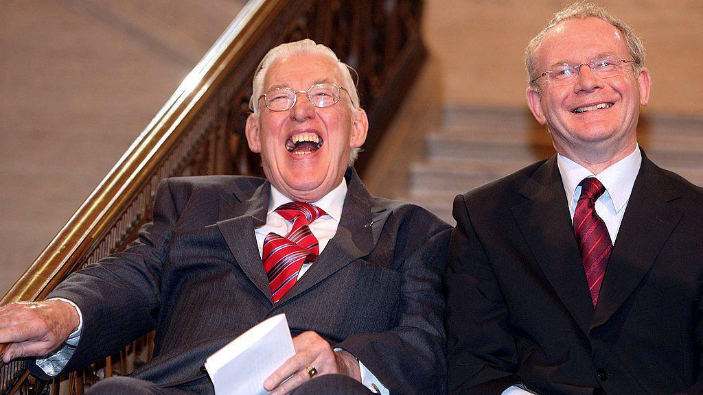 Ian Paisley (left) and Martin McGuinness sat in suits at Stormont in Belfast, both laughing.