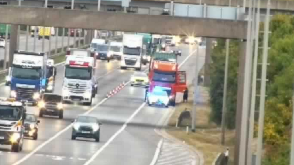 Two lanes have been closed on the motorway for a short distance. A lorry has been pulled to the side of the road and cones are blocking off on of the lanes on the motorway. Traffic is moving past the lorry and there is a police car on either side of it.