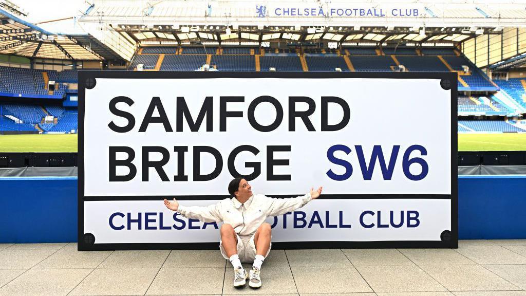 Sam Kerr sits underneath a sign of 'Samford Bridge' 