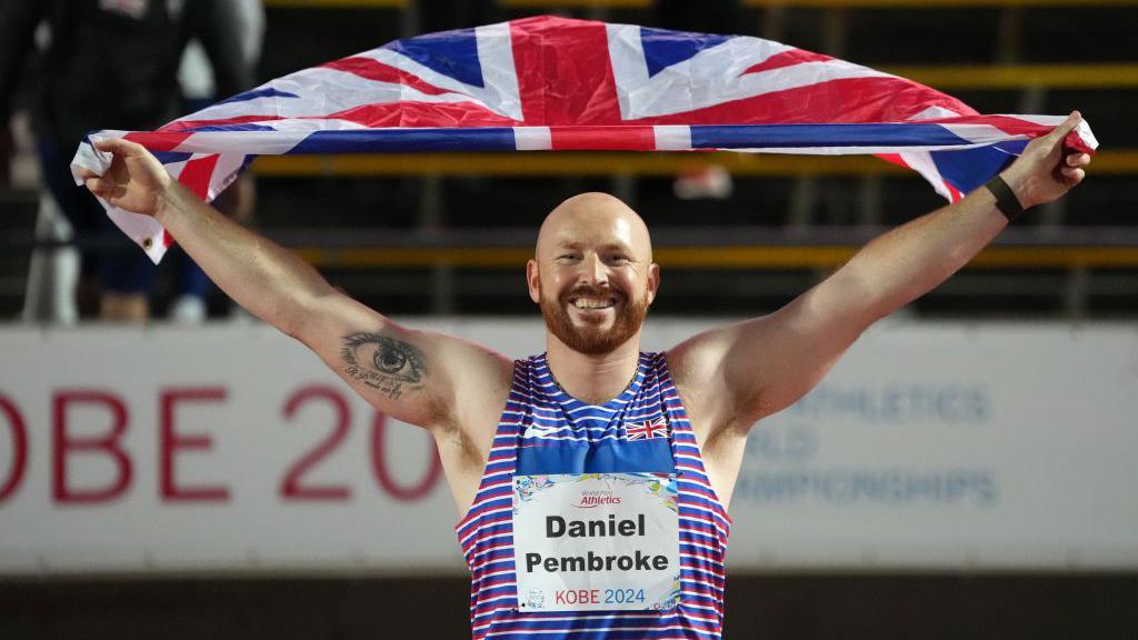 Daniel Pembroke is wearing a blue, red and white striped vest. He is bald and has auburn facial hair. He has a tattoo on his arm of a large eye. He is holding up the Great British flag above his head