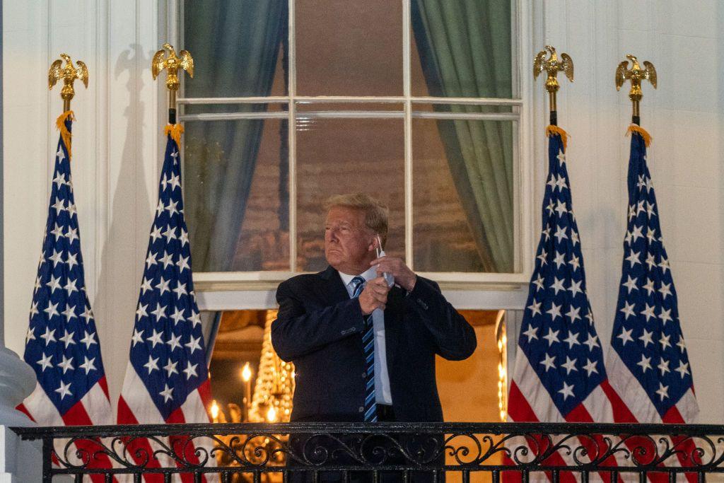 Donald Trump, in a thick, dark overcoat, removes his protective mask on the White House balcony in Washington in October 2020. Four US flags are draped behind him.