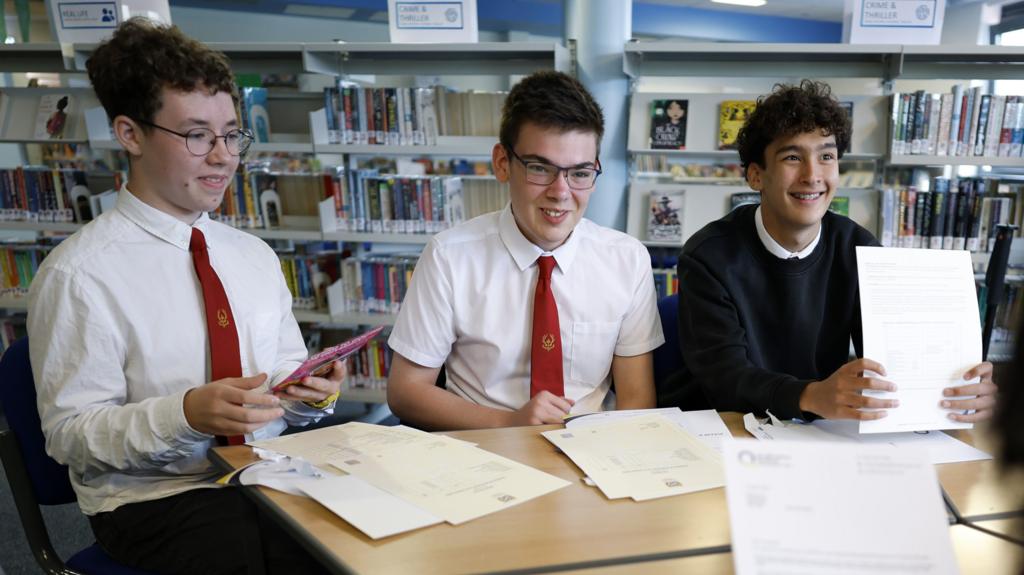 Three male students sit around a table in a library with exam results papers spread out in front of them. Two students have glasses and are wearing white shirts with red ties, while another wears a dark jumper and holds up a results sheet. Bookshelves filled with books are visible in the background.