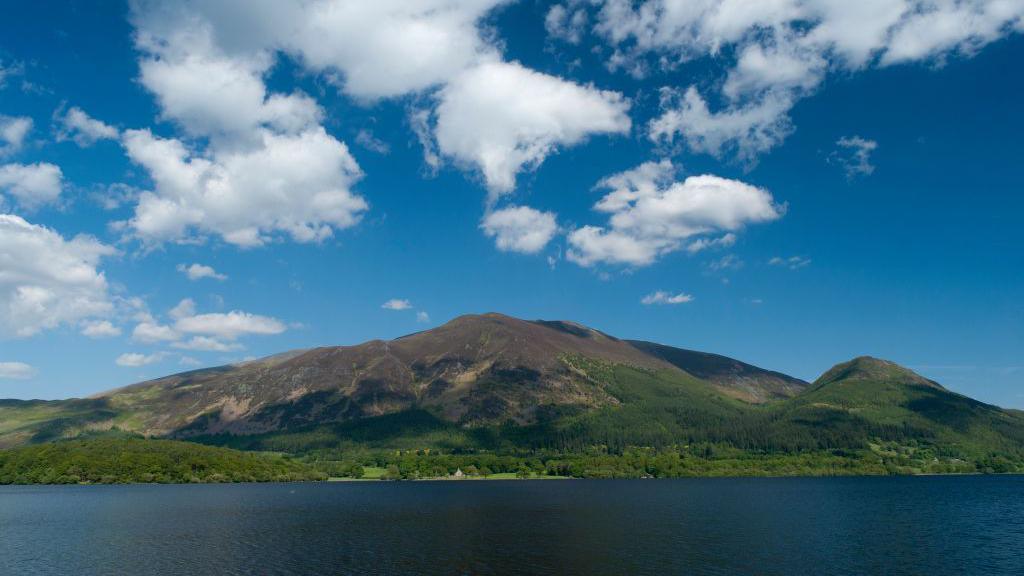 The Skiddaw range with Bassenthwaite in the foreground. Under a blue sky sits a green and brown wide triangular mountain with a dark blue lake in the foreground.