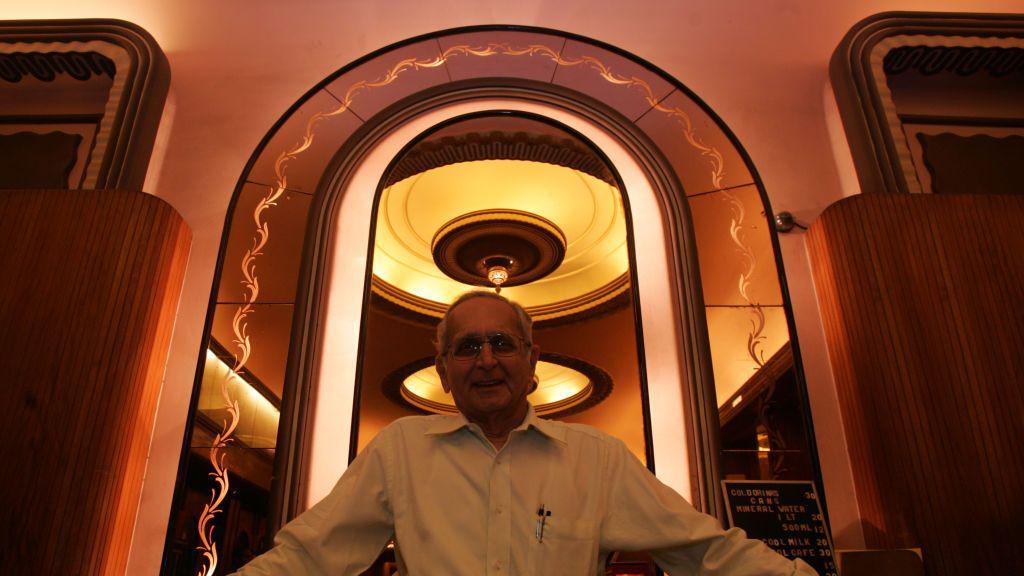 Nazir Hoosein, owner of Liberty Cinema, pictured smiling into the camera. His figure is framed by an arch, with circular Art Deco lights also behind him.
