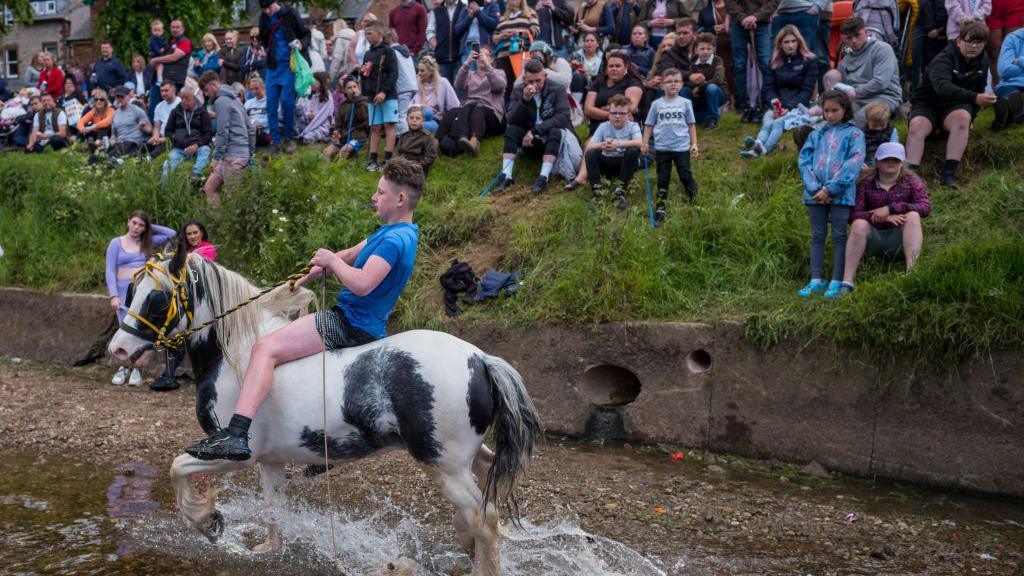 A teenager riding his horse into the River Eden, while a crowd of people watch from the bank on 8 June 2024 at the Appleby Horse Fair.