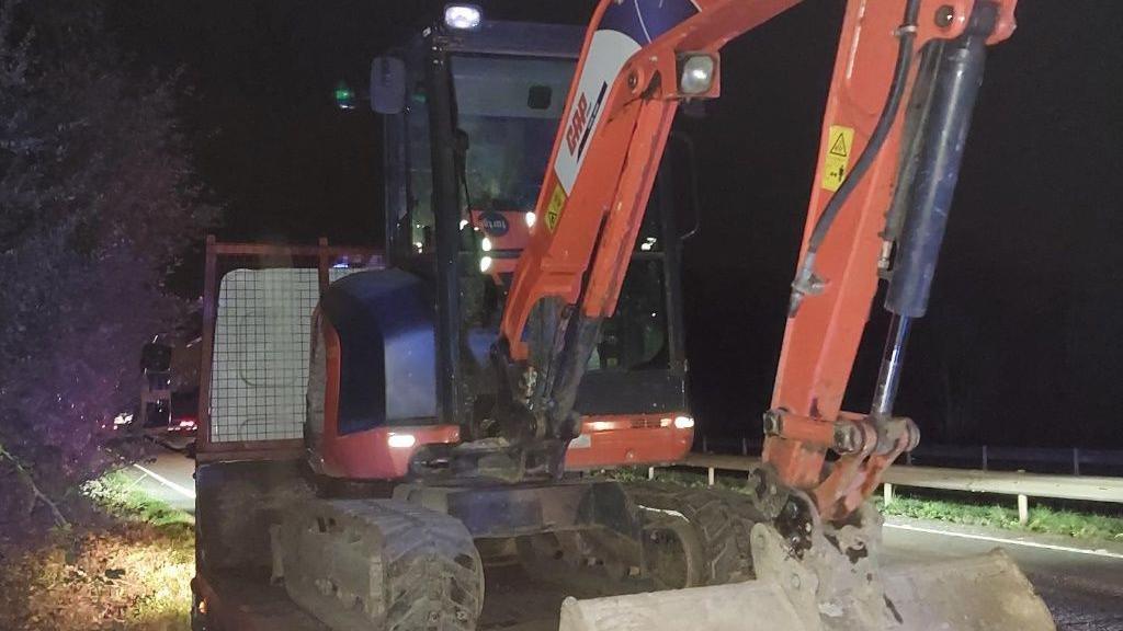 The image shows a stolen orange digger on the back of a trailer car at night. The sky is black and the trailer is parked at the side of a road. The headlight of the trailer car have been left on and the digger is lit up by headlights from another vehicle not in the picture. 