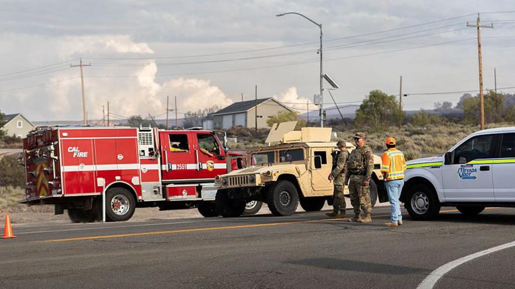 Emergency responders stand near vehicles as smoke from wildfire rises in the background.