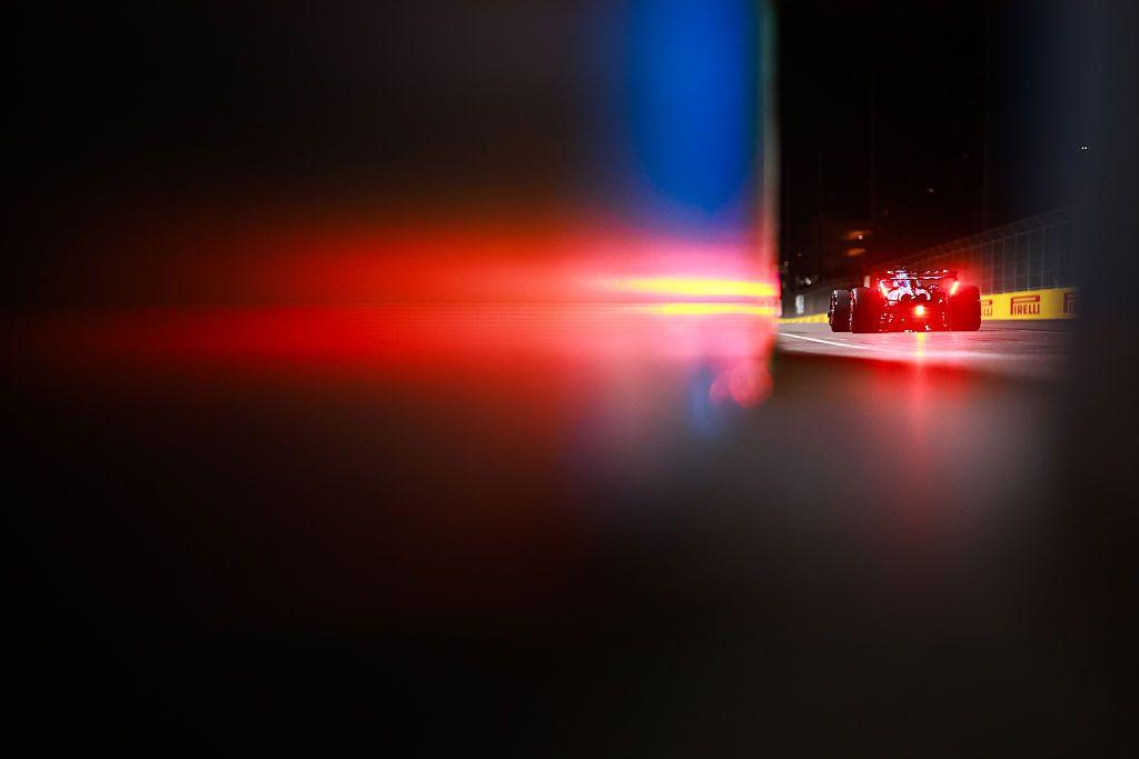 A Formula 1 car is seen from behind on a dimly lit track at night, with bright red taillights glowing and reflecting off a blurred surface in the foreground, creating a dramatic, high-speed atmosphere.