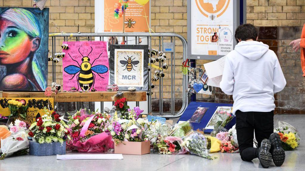 A young man kneels in front of the floral memorial at Victoria station during the commemoration. Colourful flowers, posters and pictures of Manchester bee icons are shown.
