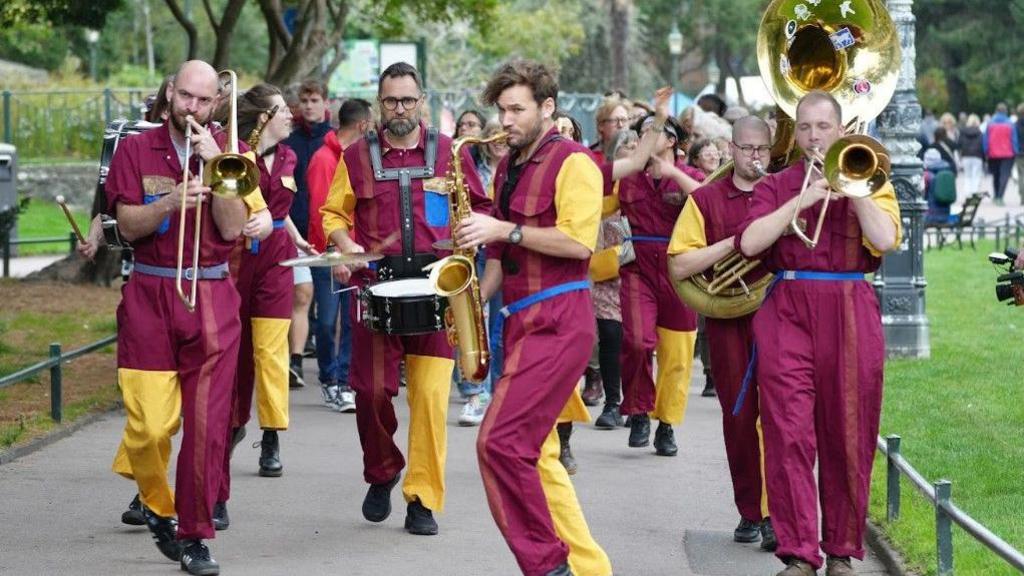 Brass band walking towards the camera playing their instruments wearing dark purple and yellow costumes.