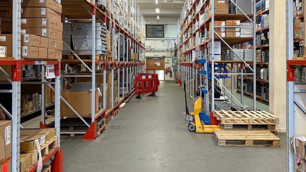 A large hall with a grey floor and wood shelving in a metal frame. The shelves are full of brown and white boxes of various sizes.  There is a white wall at the back with a control room window on the first floor.