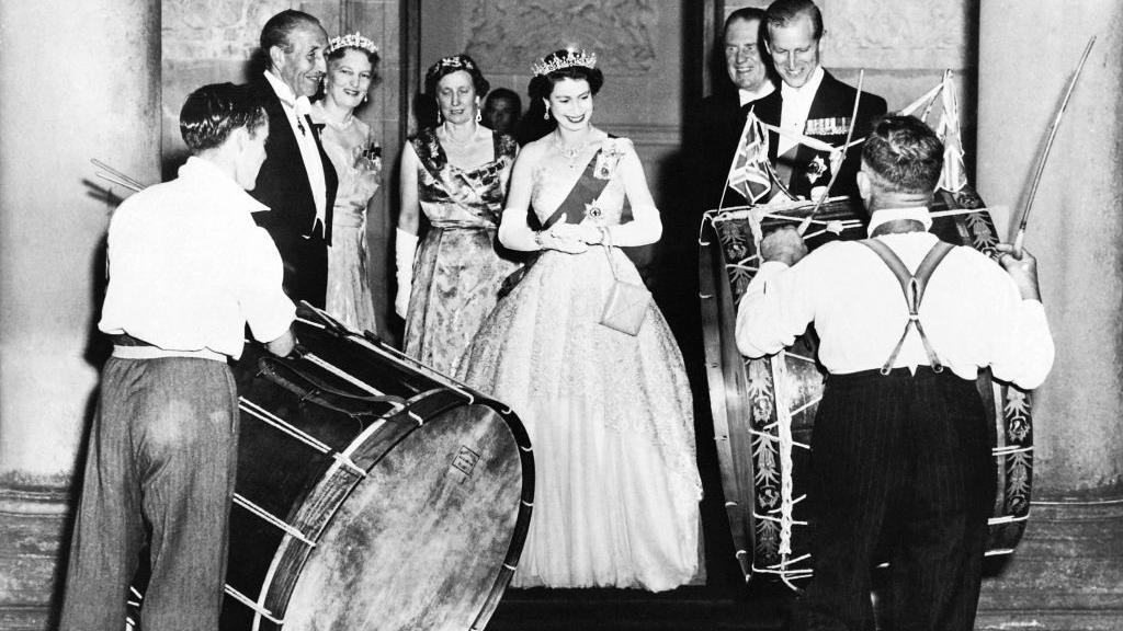 A black and white archive photo showing Queen Elizabeth II in a formal ballgown standing in the centre of the photo. In front of her are two men in white shirts playing Lambeg drums. To her left is her husband Prince Philip who is wearing white tie. 