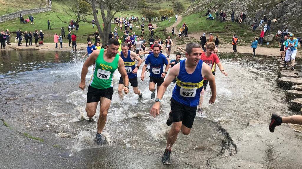 Crowds of runners racing through water, behind them is grass and a dry stone wall. 