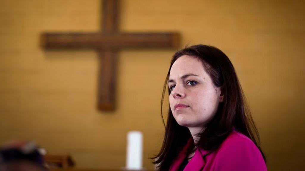 Kate Forbes looks seriously off to the side, standing in a church, a wooden cross behind her and a white candle below it. She wears a pink blazer.