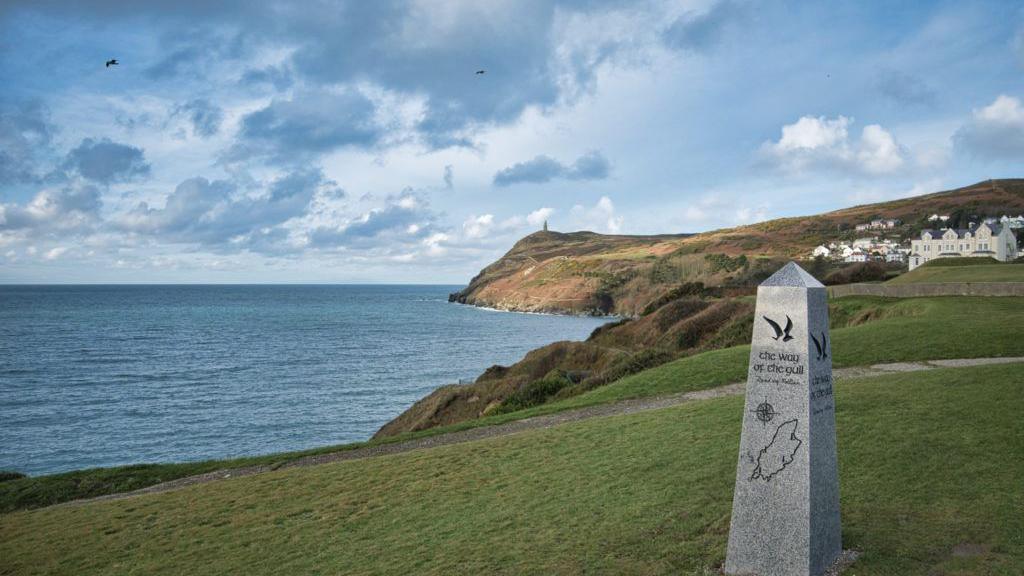 The coastline in Port Erin with Bradda Head in the background behind a tall stone Raad ny Foillan marker, which has a map of the island, a gull and a compass engraved in it.