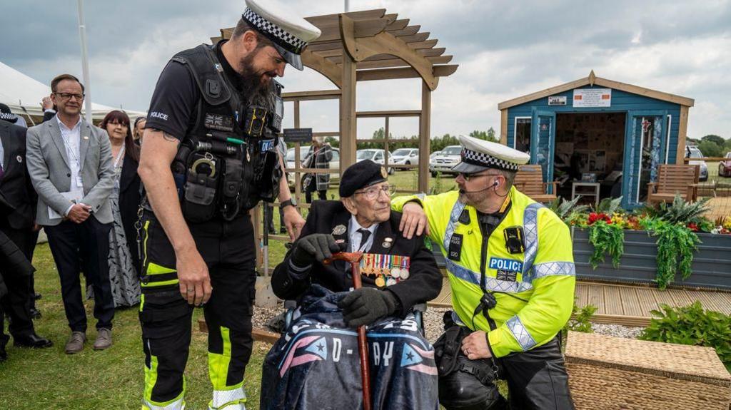 Two police officers stand beside Don Sheppard, who is sitting down outside at the memorial