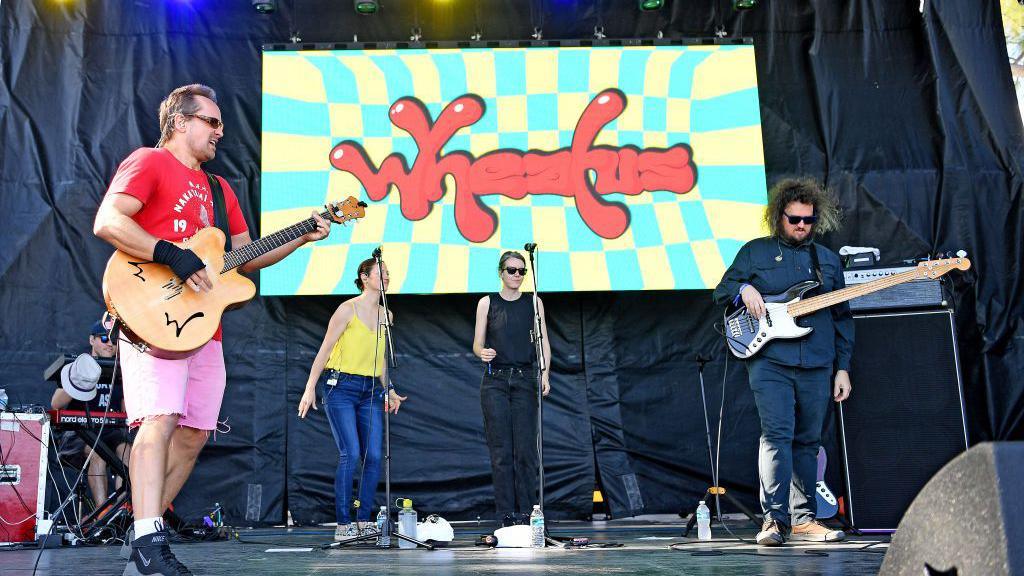 Wheatus performing on stage. Two women with dark hair, one with glasses with a man with long hair and glasses and a man with big dark hair and glasses. The men have guitars. The woman are singing. It says Wheatus on a screen in the background.