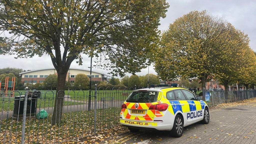 Bede Park - a leaf-strewn pathway, with a police car parked by railings and a grassed playing field