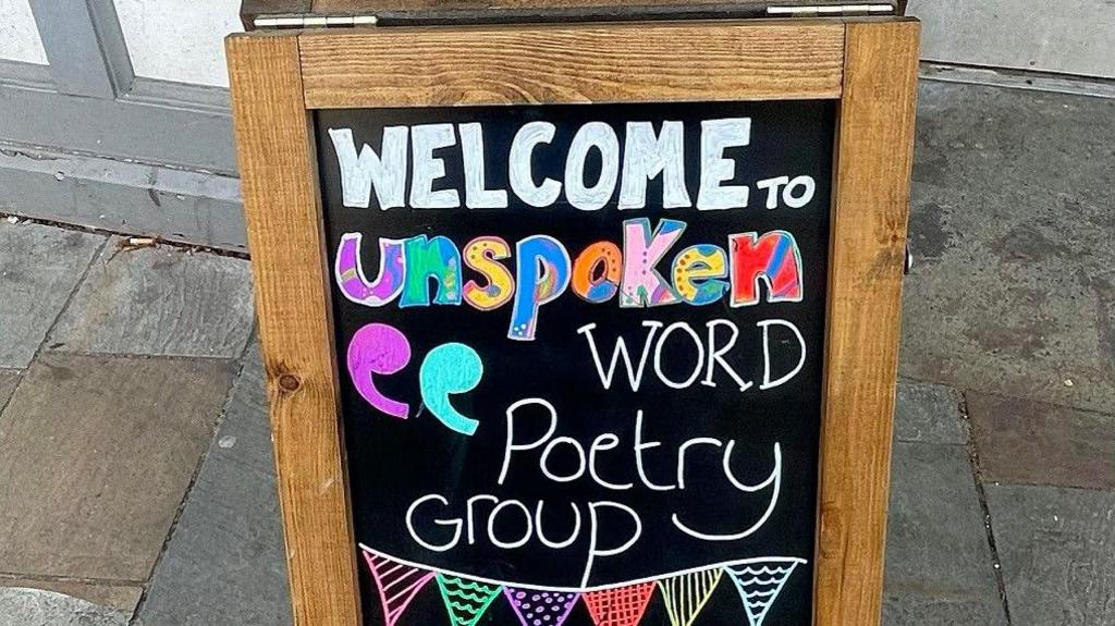 A blackboard with the words 'Welcome to Unspoken Word Poetry Group' written on it. The word unspoken has been written in colourful letters.