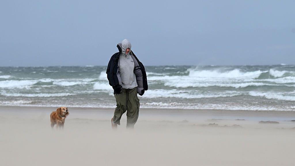 Man walking along beach with dog in wind. Rough looking sea behind.