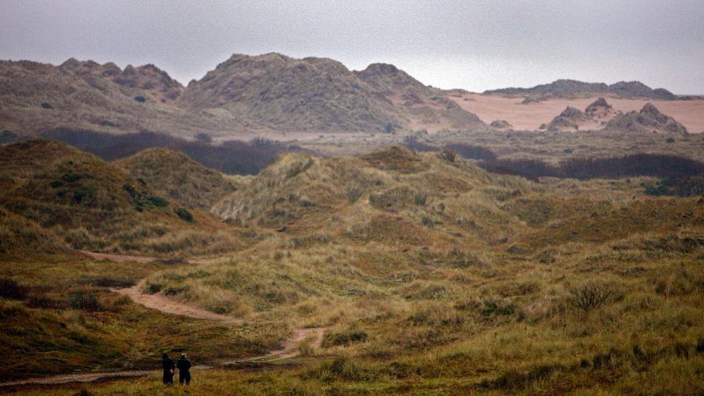 A wide view of an area of large sand dunes and grass-covered sand. Two people are standing in the foreground, dwarfed by the landscape. It is greay and overcast.