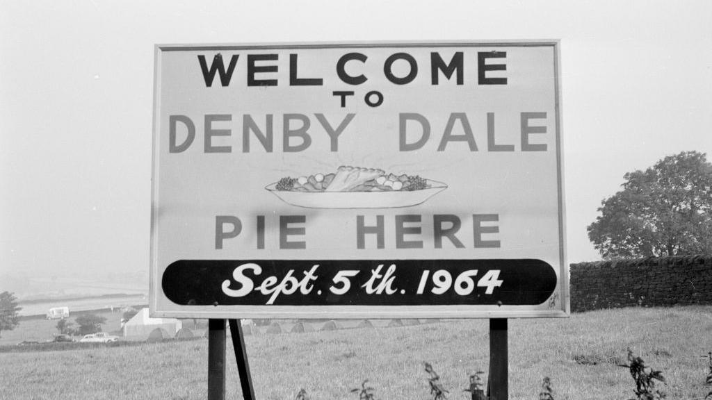 A black and white photograph of a sign that reads: 'Welcome to Denby Dale Pie Here Sept. 5th 1964'
