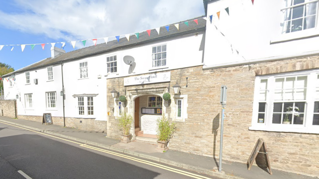 A pub which also has a section of brown brick wall. There is a white sign on the building that says "The Swan Hotel". There is colourful bunting attached to the side of the pub, which stretches across the road. The pub sits next to the road.