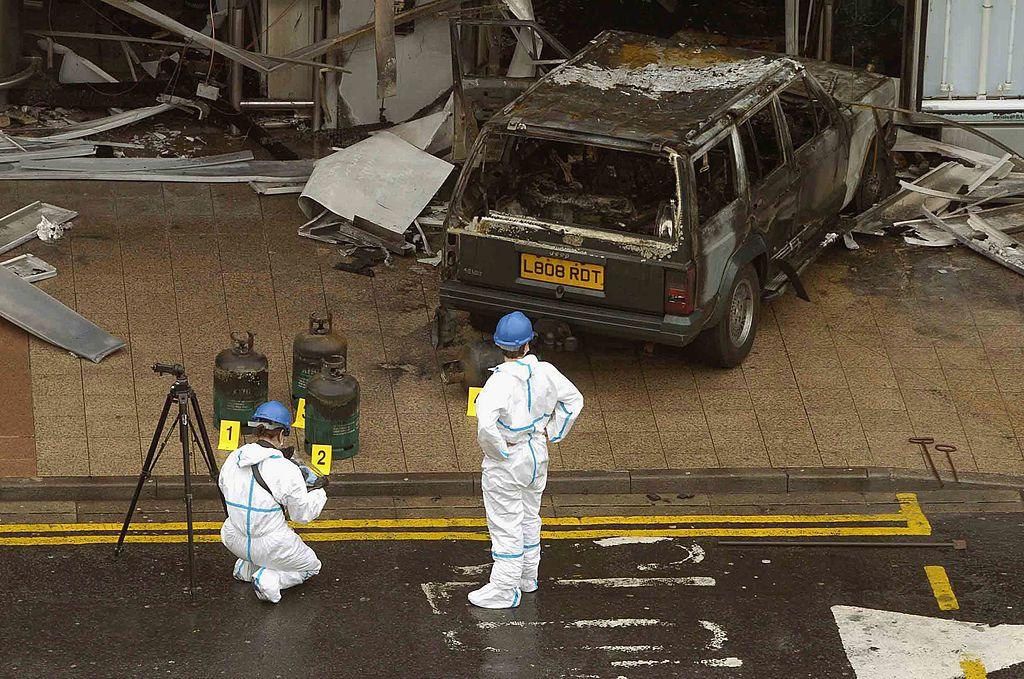 Forensic experts examine the scene at Glasgow Airport terror attack. Two experts in white suits are pictured looking at charred gas cannisters and a burnt out jeep