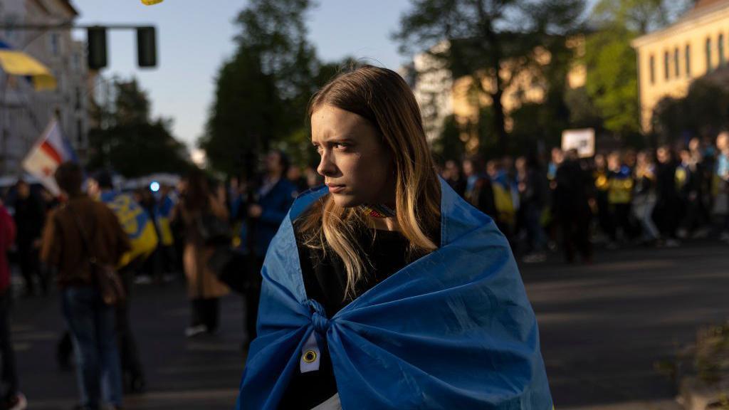 A woman with blonde hair, draped in a blue Ukraine flag, looks to her side sadly at a protest in Berlin to commemorate the millions of Ukrainians who died during World War II on VE Day in 2023.