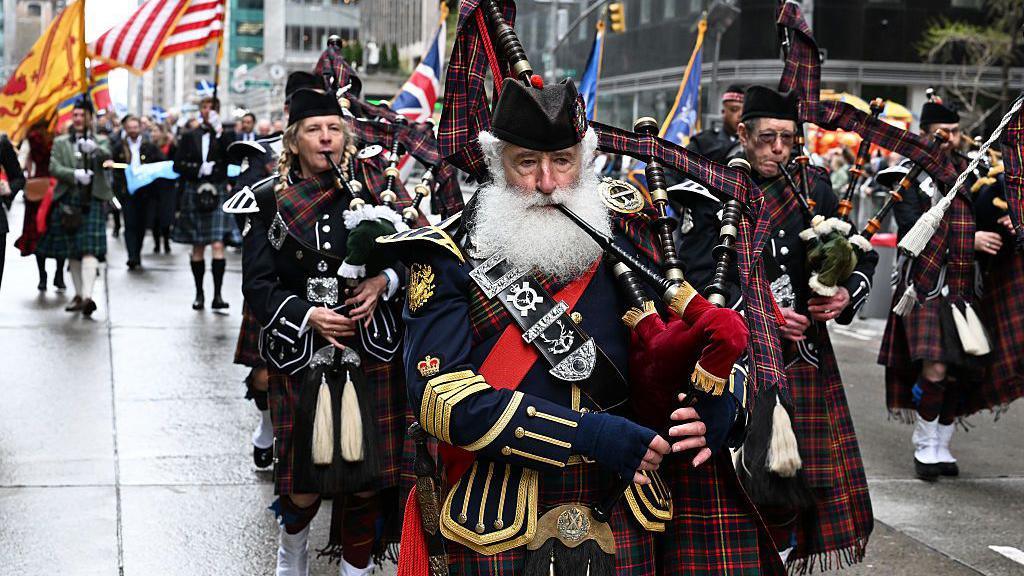 An impressively white-bearded man is playing the bagpipes and leading a band all in full Highland dress down the streets of New York. Everyone appears to be having a nice time.
