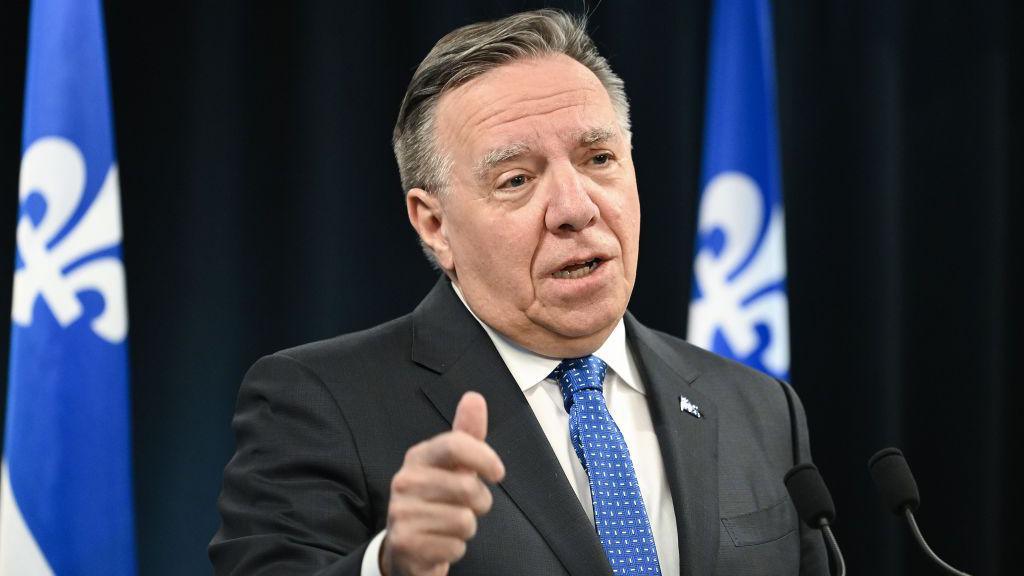 Quebec Premier Francois Legault, wearing a dark suit and a blue and white patterned tie, speaks into two microphones. Behind him, against a black backdrop, are two blue and white Quebec flags. 