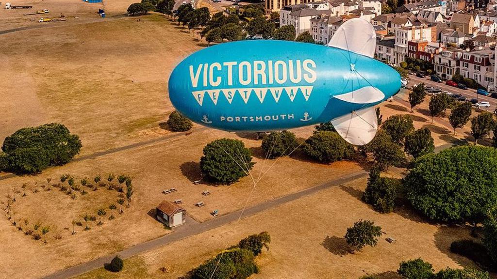 Teal coloured large balloon floating above parched ground below and buildings seen in the distance.