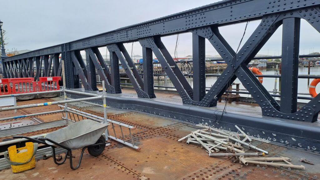 The work site on Corporation Road Bridge. The steel structure has been freshly-painted. A wheelbarrow, scaffolding, a ladder and barriers are visible. 