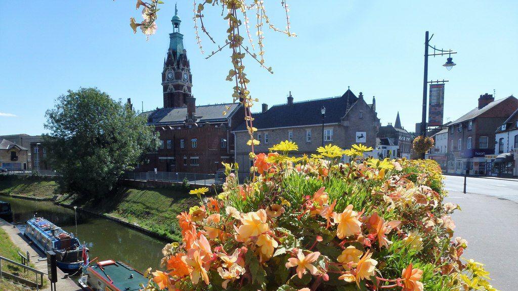 Flowers on a road bridge over a river with some boats moored up on the left, a large ornate clock tower can be seen behind with a statue of Britannia in green bronze on top.